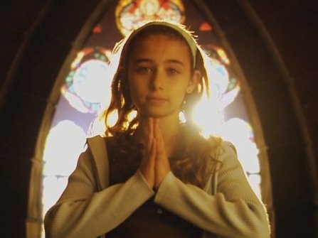 A little girl in front of a stained glass window with her hands together in prayer